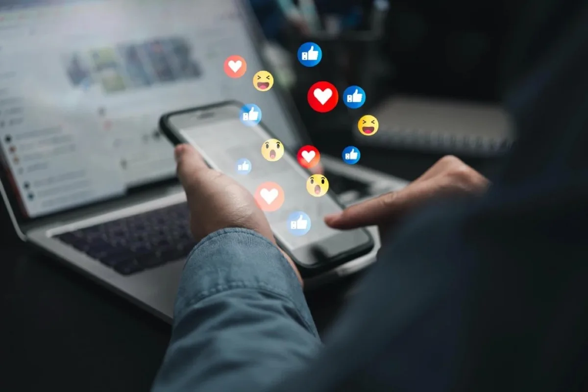Person bedient ein Smartphone, während Social-Media-Reaktionen wie Daumen hoch, Herzen und Emojis vom Bildschirm aufsteigen