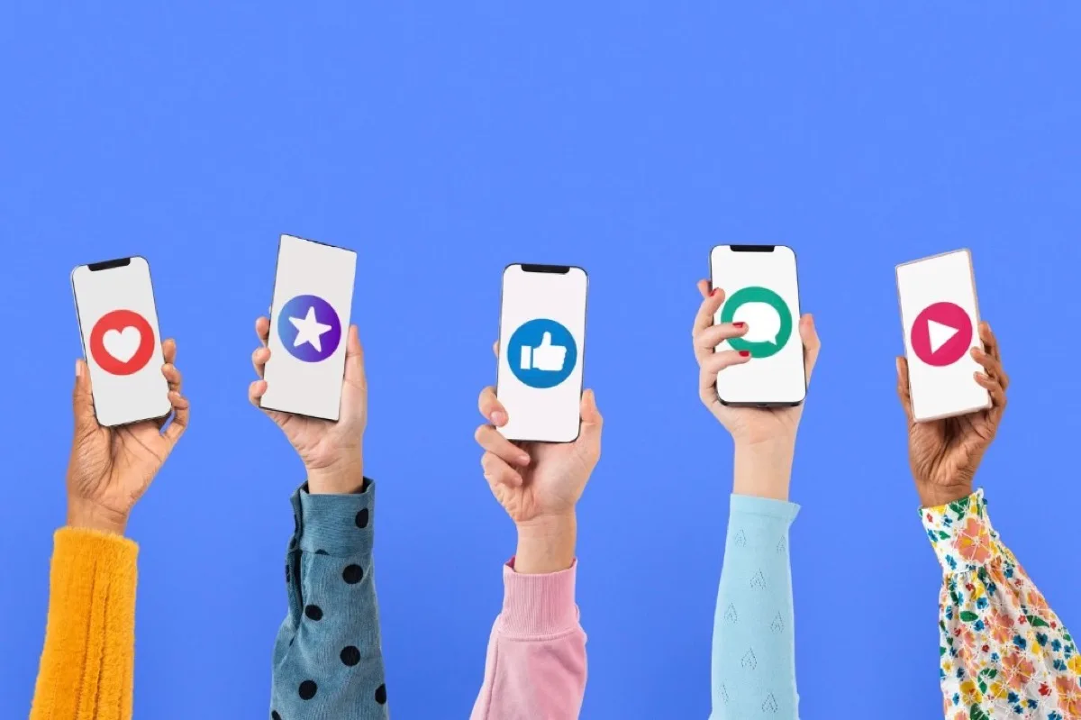 Fünf Hände halten Smartphones mit verschiedenen Social-Media-Symbolen wie Herz, Daumen hoch und Play-Button vor blauem Hintergrund